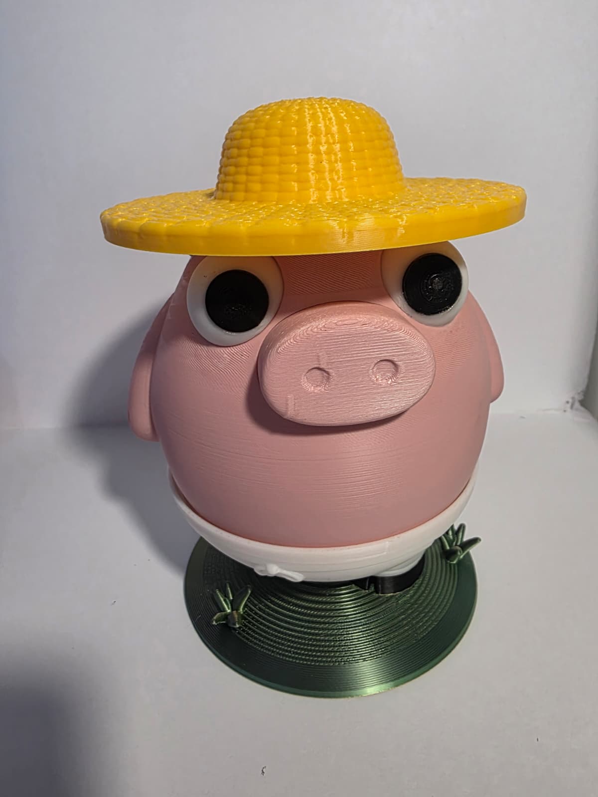 A 3D-printed pig character figurine wearing a yellow corn-cob-style hat, standing on a green circular base against a plain white background.
